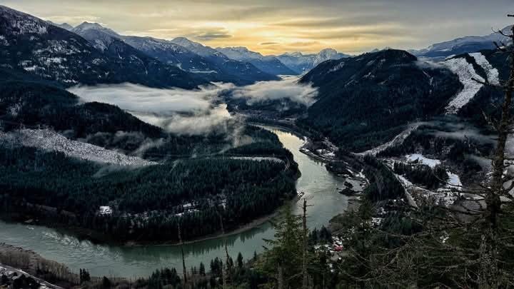A sweeping mountain valley in winter, with a winding river reflecting soft golden light beneath an overcast sky. Snow-dusted peaks rise dramatically around dense evergreen forests, while low clouds drift above the treetops, adding depth and serenity to the scene. The contrast between dark green trees, white snow, and the glowing river creates a tranquil, cinematic atmosphere.