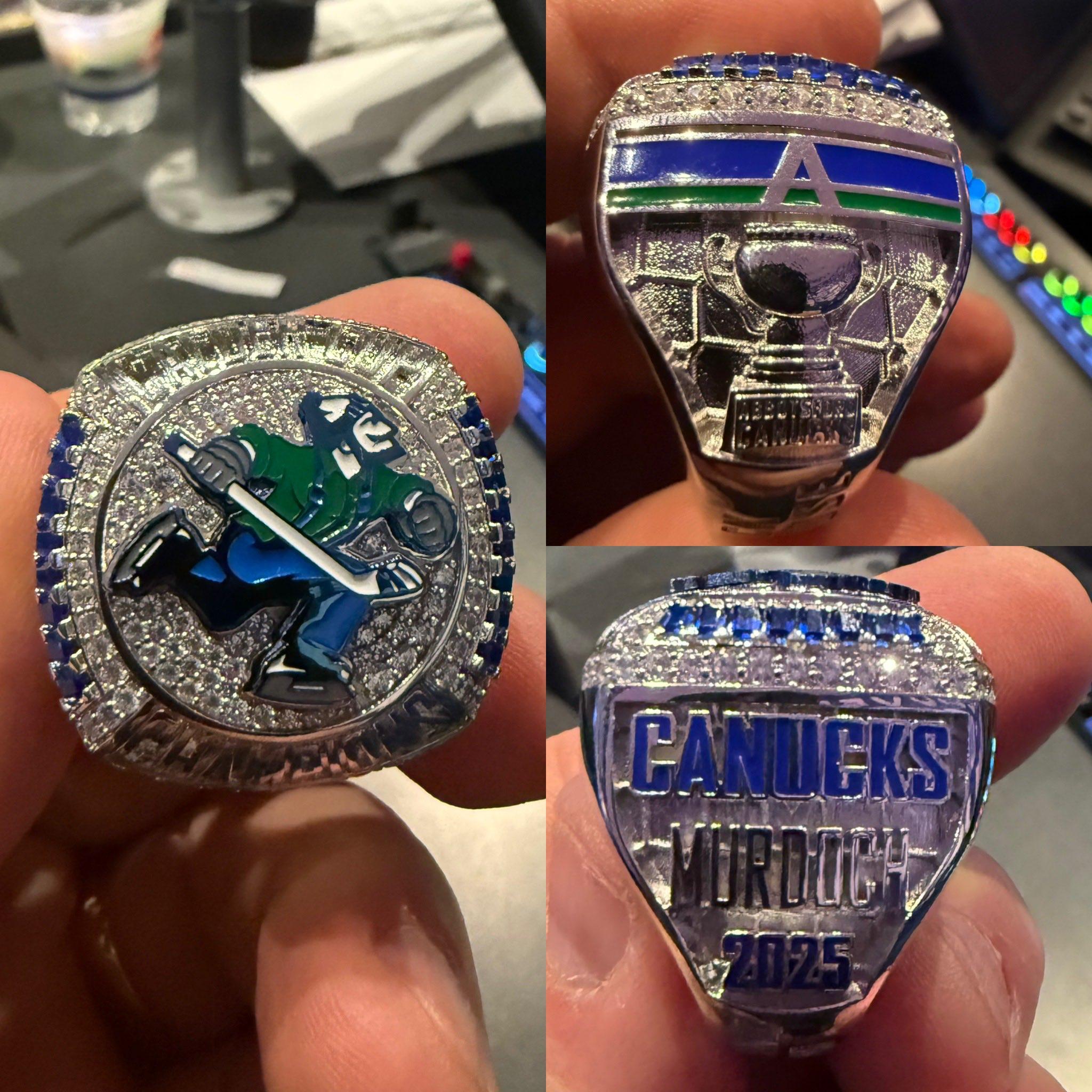 A championship-style hockey ring shown from three angles. The front face features a stylized hockey player in green and blue gear skating with a stick, set against a background of small sparkling stones, with the words“CALDER CUP CHAMPION” engraved along the bottom and top edge. One side of the ring displays a trophy above the words “ABBOTSFORD CANUCKS” and a blue‑and‑green “A” logo. The opposite side reads “CANUCKS” in bold blue letters, with the engraved name “MURDOCH” and the year “2025” beneath it.