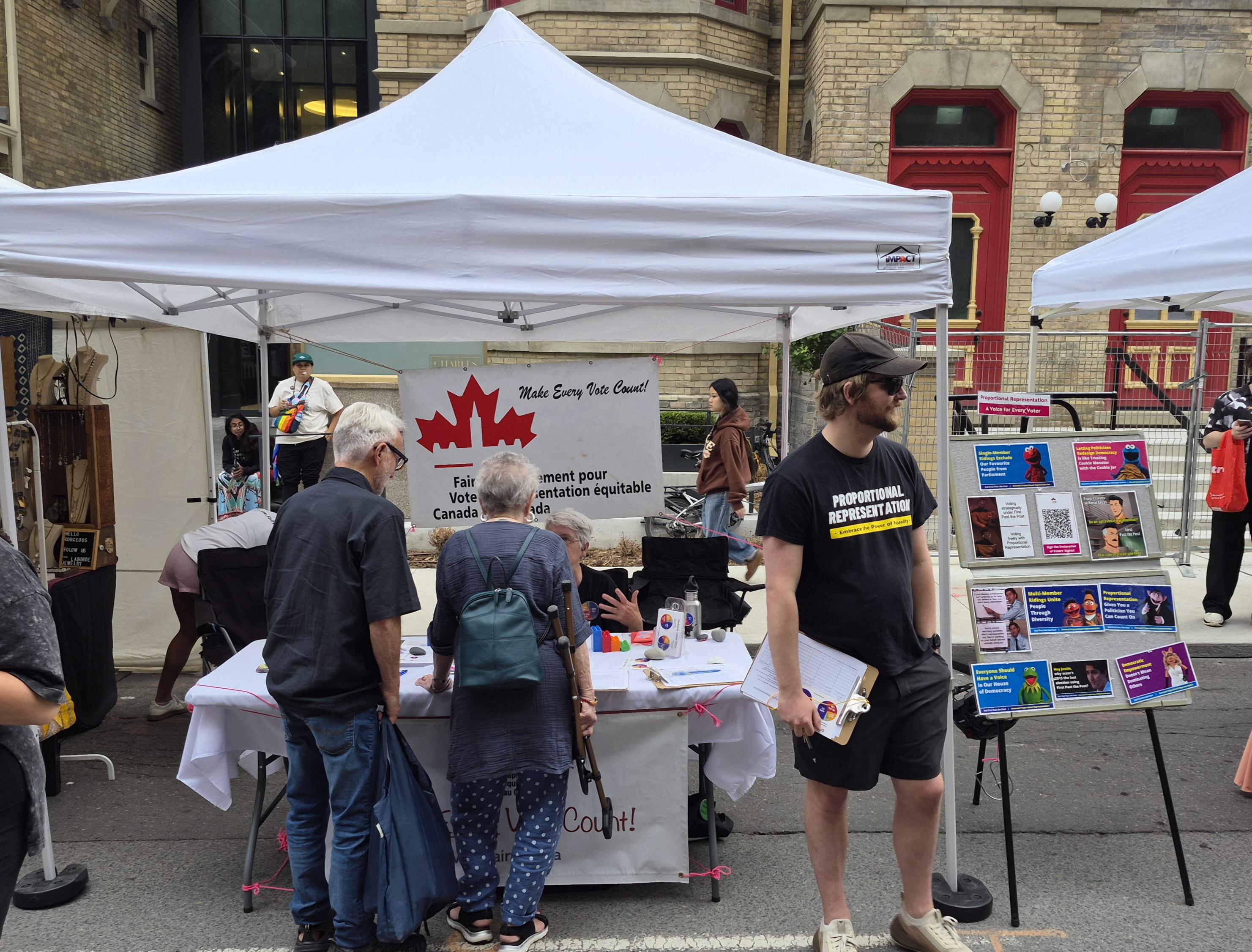 Picture of a white tent top on four posts, with a table underneath. The table has a white tablecloth and brochures, cards, and stickers.
Two festival goers are signing a form, one volunteer is seated behind the table talking to them. Another volunteer in sunglasses stands next to a board full of memes. One meme shows Ms Piggy and says Democratic Empowerment doesn't mean dominating others. Another meme shows Burt and Ernie and says Multi Member ridings United people through diversity. Another shows Kermit and says everyone should have a voice in our house of democracy. Another shows The Count and says Proportional Representation gives you a politician you can count on. Another shows the cookie monster and says letting politicians redesign democracy is like trusting the cookie monster with the cookie  jar. Another shows Elmo and says Single Member ridings exclude our favourite people from parliament.