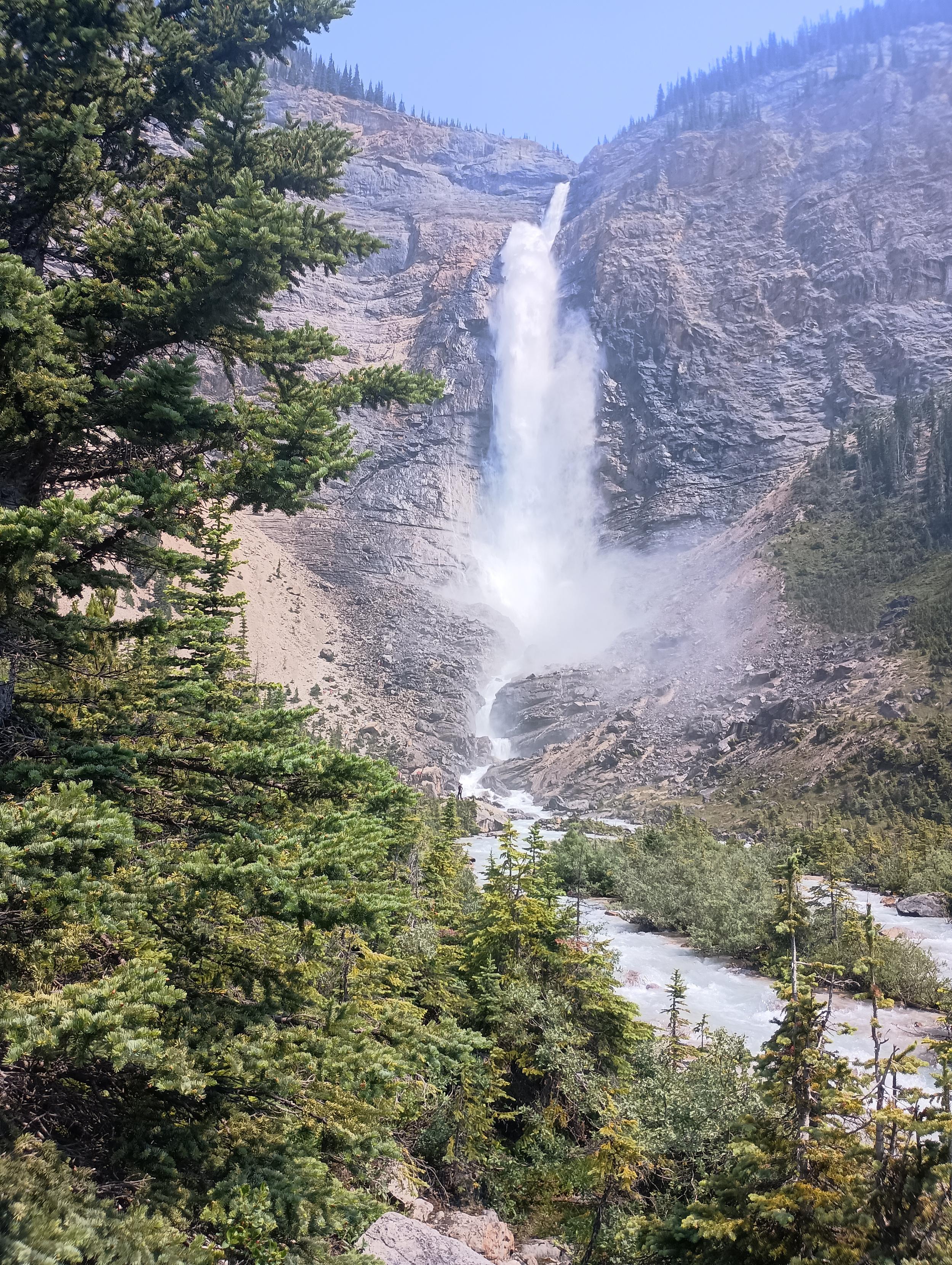 A thin ribbon of water emerges over the edge of a mountain cliff, and falls 18 m before ramping over another ledge, like water going over a ski jump.

As the water plummets the rest of the way down the mountain, it bounces off the cliff face, spreads out and generates a lot of spray that blows off to the right.