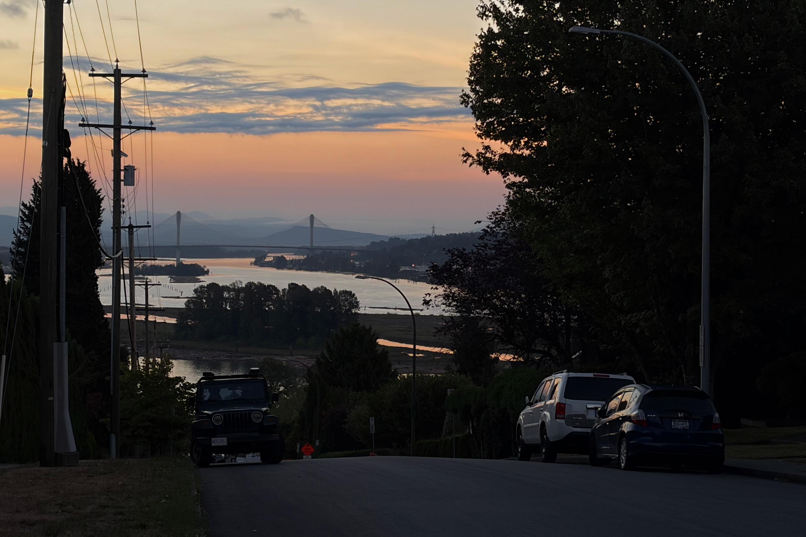 A road with a Jeep and two cars parked on the side, overlooking a river with a bridge in the distance during sunrise.