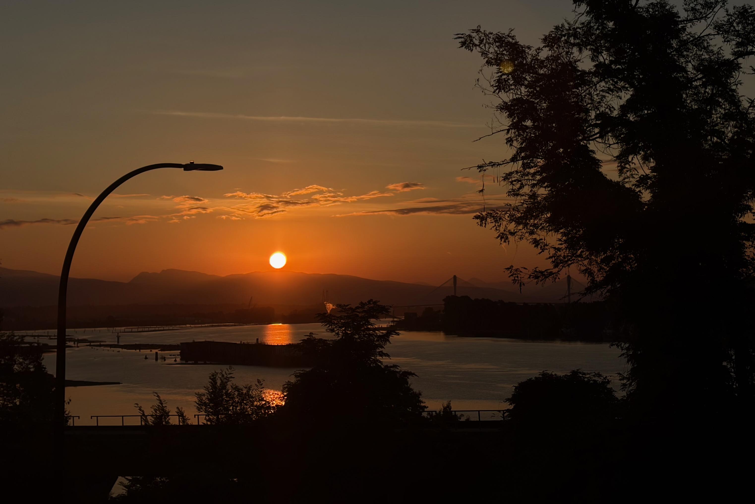 A sunrise view over a river with a bridge in the distance, trees, and a streetlight on the left. The sky is orange and pink, and the water reflects the colours.
