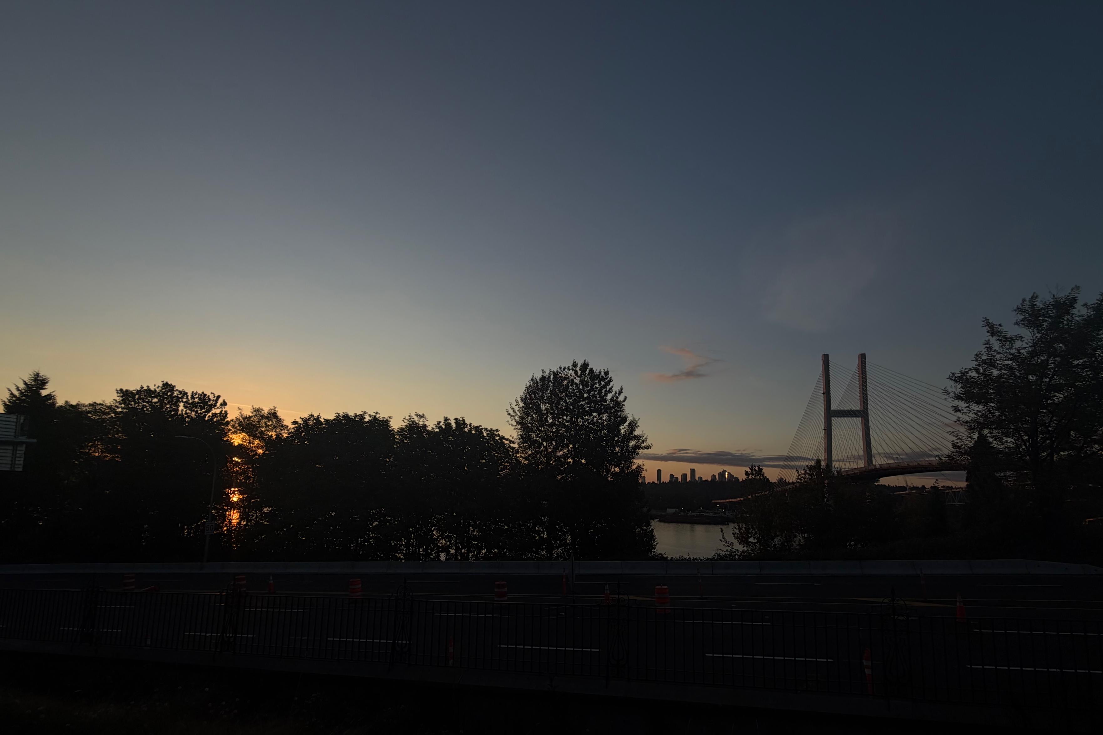A silhouetted view of a bridge over a river at sunrise, with trees and a city skyline in the distance, captured from a road with traffic cones and barriers.