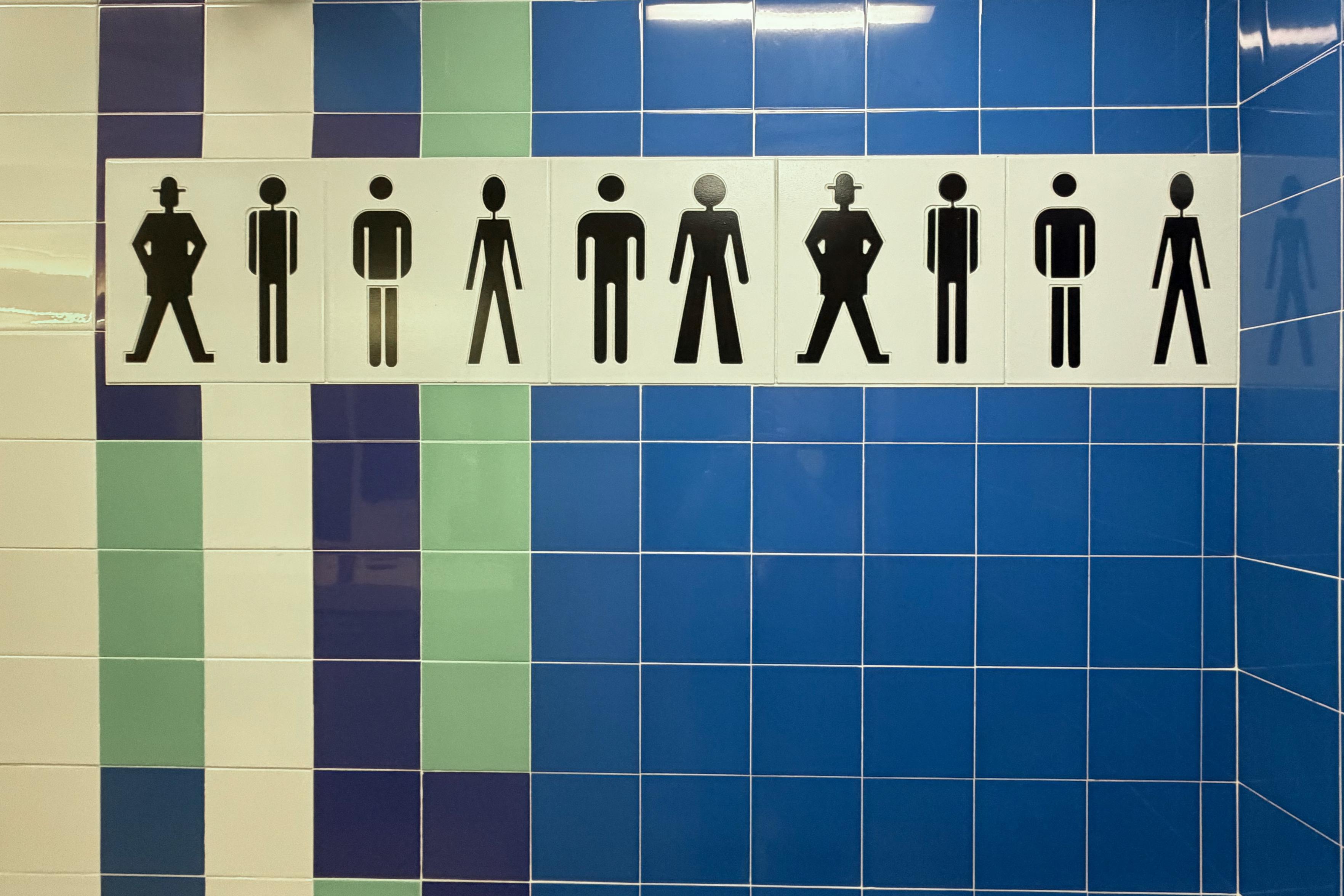 A blue and green tiled wall with a white sign displaying black silhouettes of men in various poses, likely indicating a men's restroom.