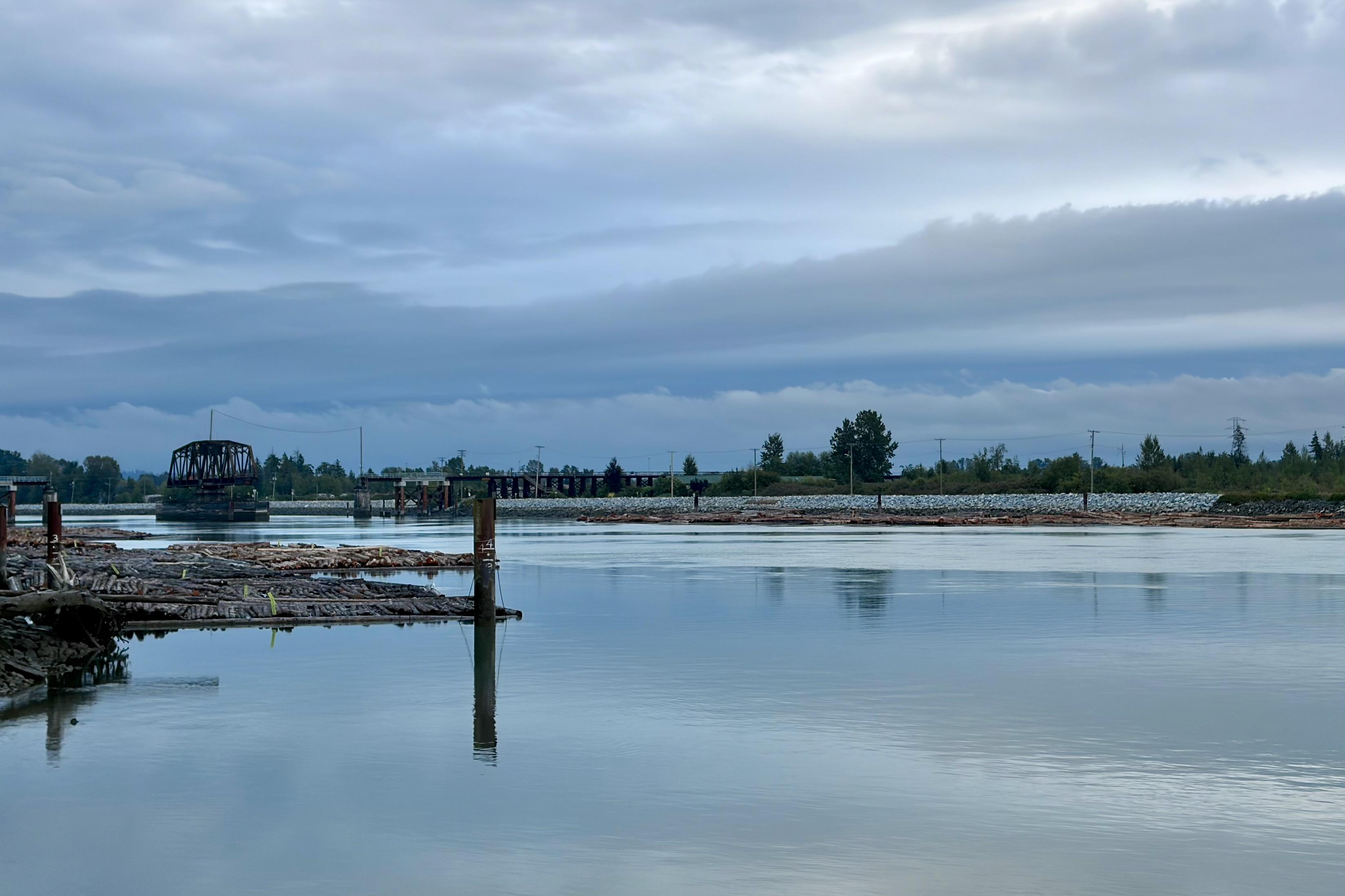 A calm river with a bridge in the distance, surrounded by trees and a cloudy sky.