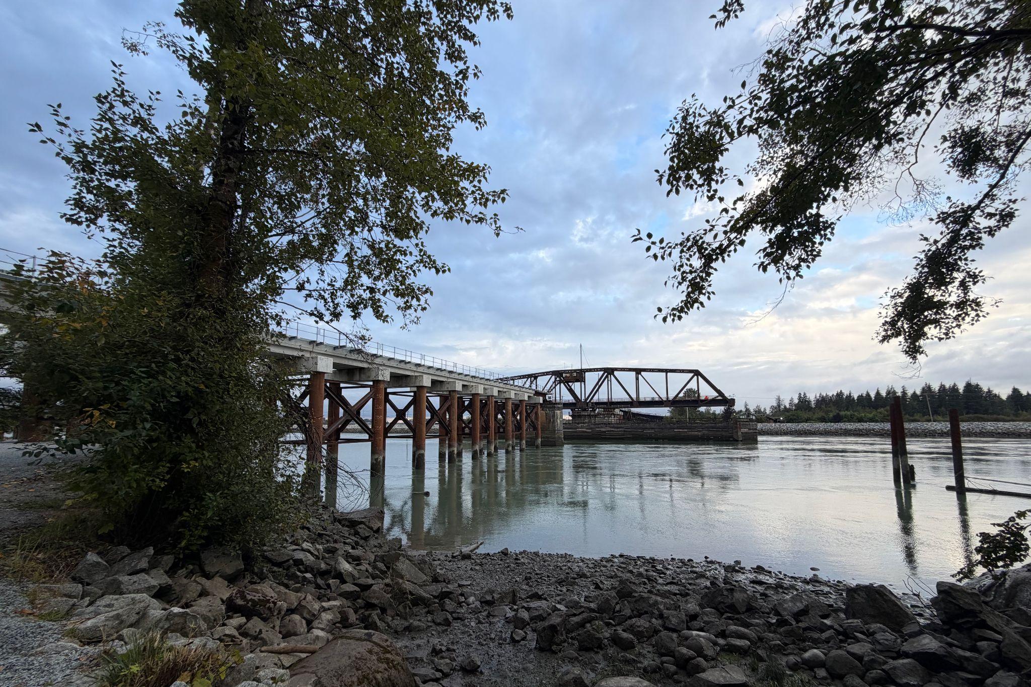 A bridge with a metal structure spans a river, surrounded by trees and rocks, under a cloudy sky.