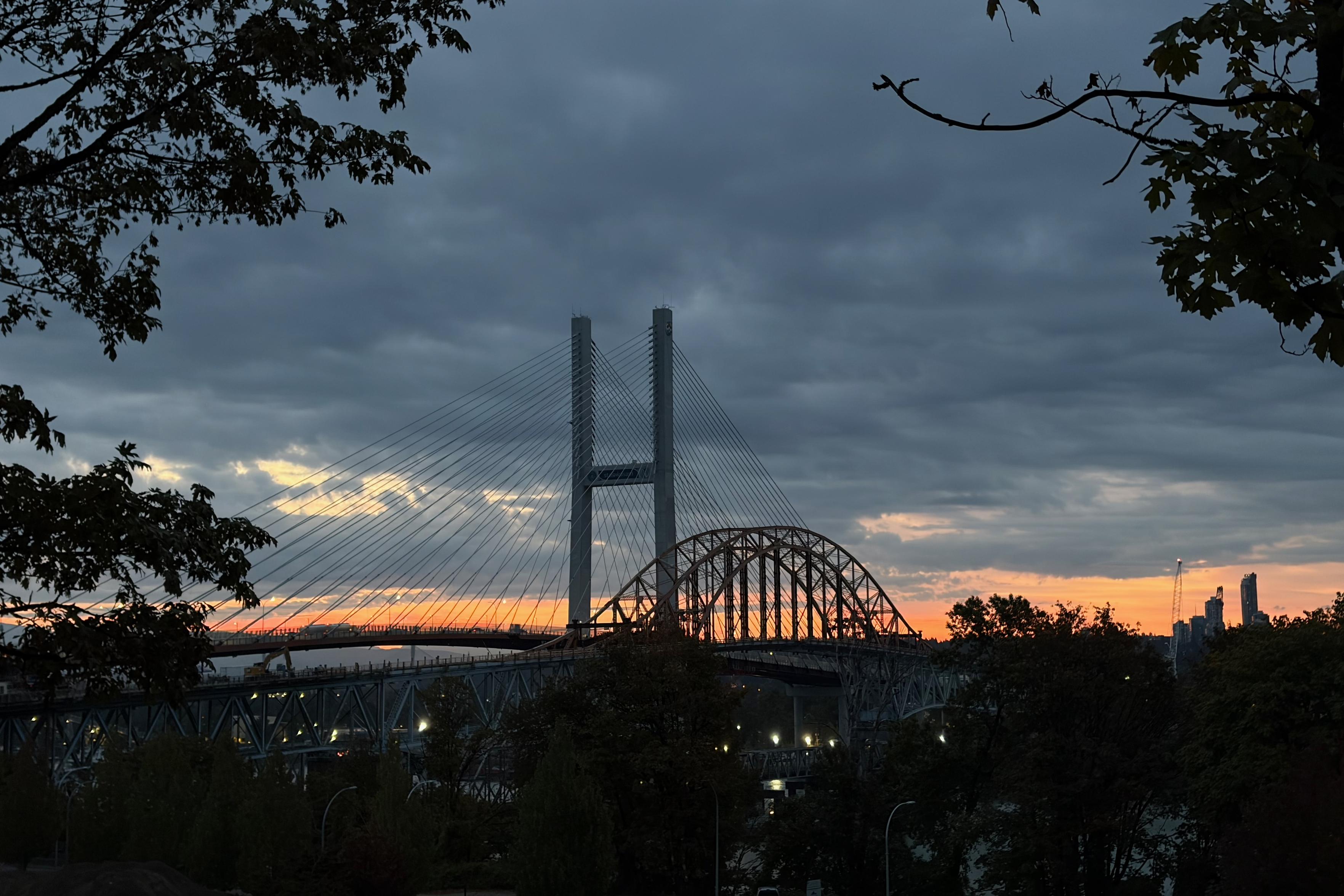 A bridge with two tall towers and a curved arch, surrounded by trees, under a cloudy sky with a sunrise in the background.