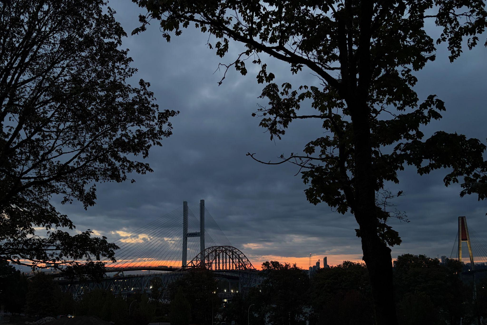 A silhouetted view of a bridge with a sunrise in the background, framed by trees and a cloudy sky.