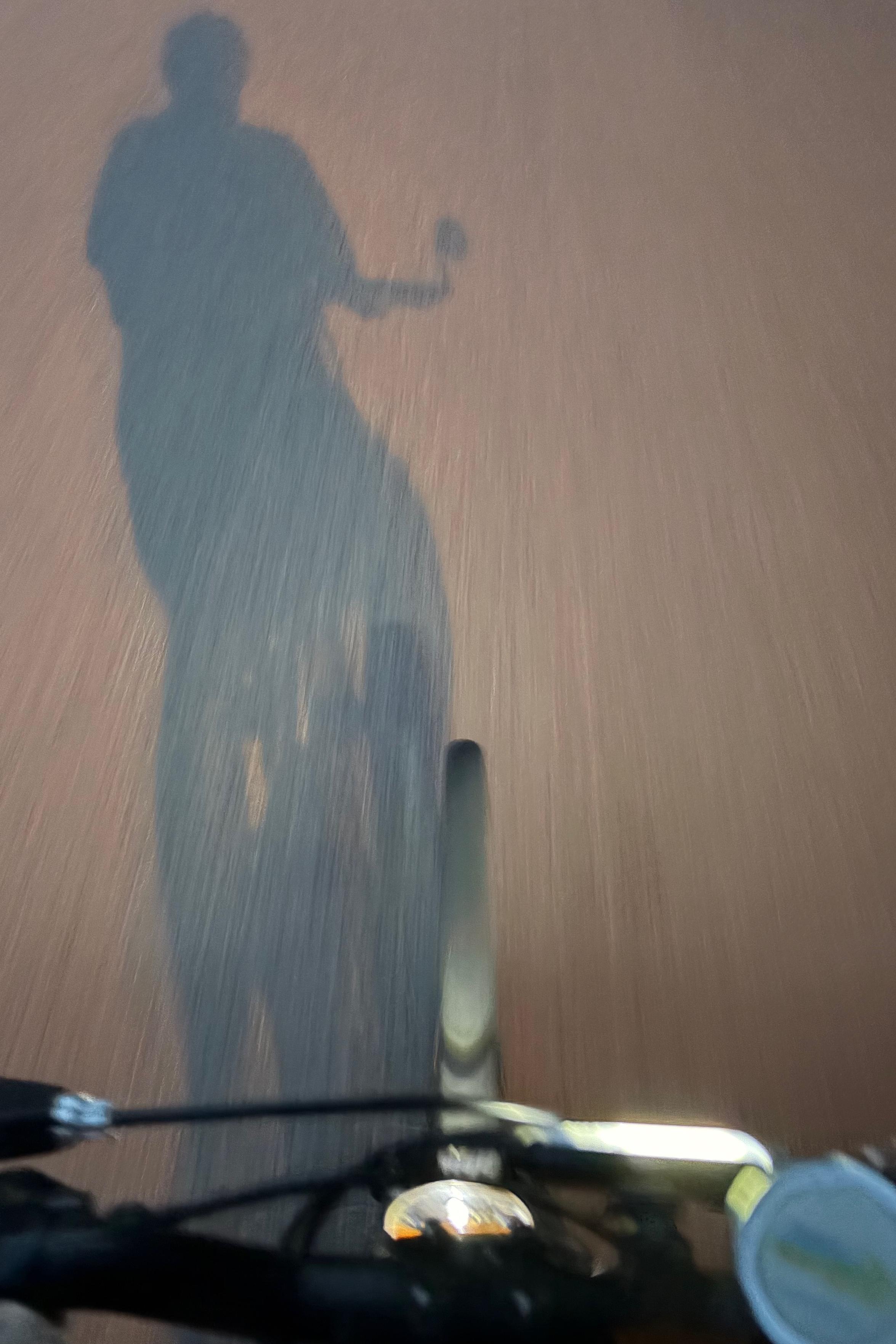 A silhouette of a person riding a bicycle on a road, with the person's shadow cast on the ground, and the bicycle's handlebars,the front wheel and a lamp visible.